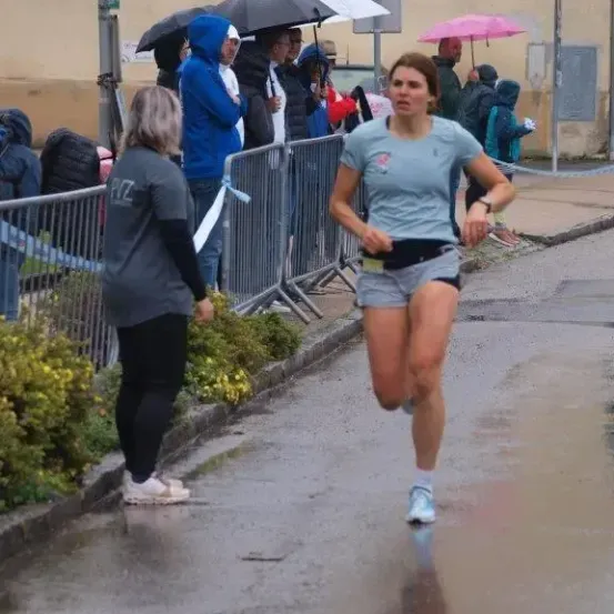 Eine Frau läuft bei Regen auf einer nassen Straße, mit Zuschauern auf dem Bürgersteig. Hinter ihr steht eine Frau in der Nähe einer Metallbarriere. Manche Leute halten Regenschirme, andere tragen Hüte und wieder andere haben Turnschuhe an.