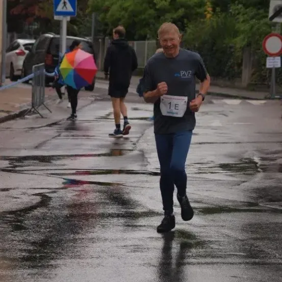 Ein Mann in einem grauen Shirt mit PVZ-Aufschrift läuft bei einem Marathon. Es gibt Leute, die am Straßenrand gehen, einer von ihnen hält einen Regenschirm.