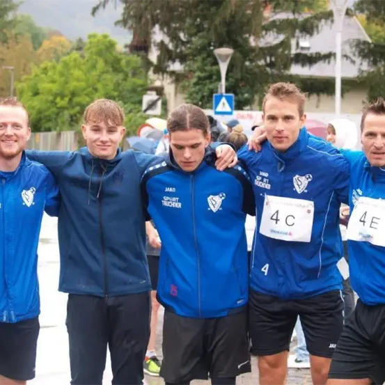Fünf Männer in blauen Jacken und schwarzen Shorts posieren für ein Foto. Sie sind draußen, wahrscheinlich bei einer Sportveranstaltung. Der Mann in der Mitte trägt ein weißes Startnummernband.
