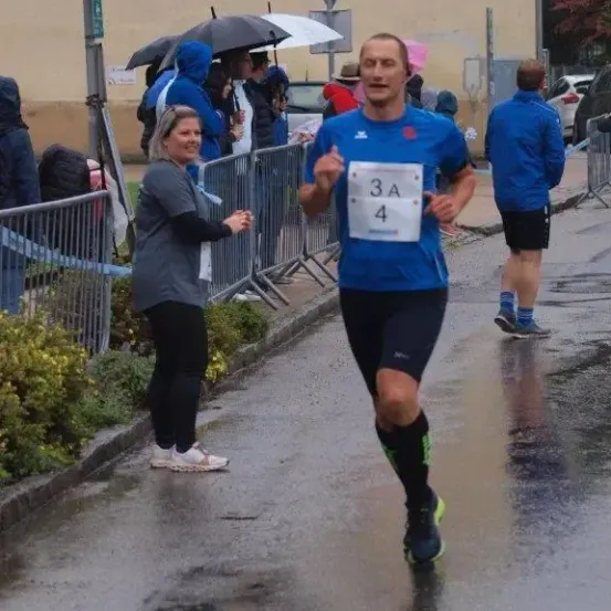 Ein Mann mit der Nummer 3A4 läuft einen Marathon, während eine Frau ihn anfeuert. Dahinter stehen Menschen mit Regenschirmen und geparkte Autos.