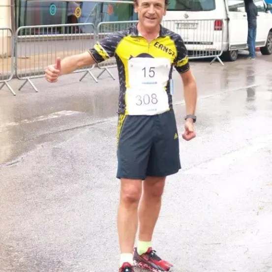 Ein Mann mit erhobenem Daumen, der ein Laufshirt trägt, steht im Regen auf der Straße. Hinter ihm ist ein weißer Van geparkt, und eine Person steht in der Nähe. Es gibt Absperrungen auf der Straße.