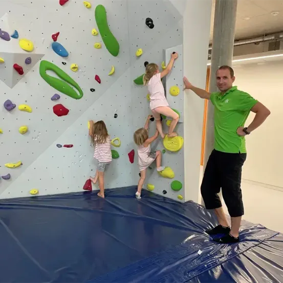 Drei junge Mädchen und ein erwachsener Mann klettern an einer bunten Kletterwand. Der Mann steht neben der Wand und unterstützt die Mädchen. Die Wand ist mit einer blauen Plane abgedeckt.
