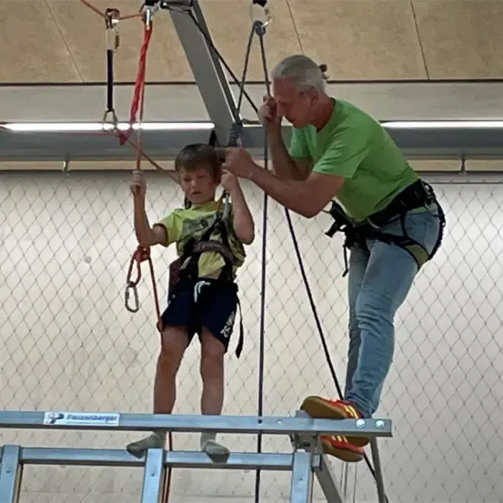 Ein älterer Mann hilft einem kleinen Jungen, beide tragen Gurte, beim Klettern an einem Gurt mit Seilen. Sie befinden sich in einem eingezäunten Bereich mit einer weißen Wand.