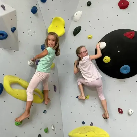 Zwei junge Mädchen klettern auf einer farbenfrohen Kletterwand, eine in einem grünen Shirt und die andere in einem rosa Shirt. Sie benutzen verschiedene Griffe und scheinen die Aktivität zu genießen.