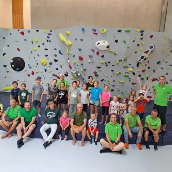 Eine Gruppe von Menschen, einschließlich Kindern, posiert vor einer Wand voller verschiedenfarbiger Klettergriffe. Einige Personen tragen grüne Shirts, während andere verschiedene Farben tragen.