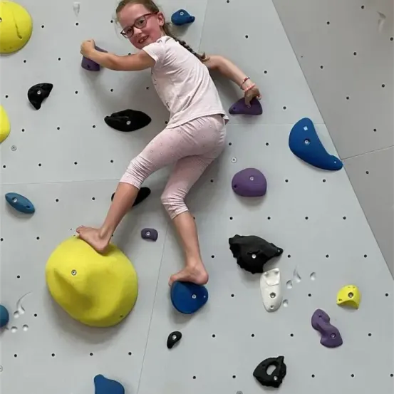 Ein junges Mädchen mit Brille klettert an einer Wand mit verschiedenen farbigen Griffen. Sie trägt ein rosa Shirt und rosa Hosen. Sie hält sich mit der rechten Hand an einem gelben Griff fest.