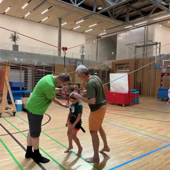 Zwei Männer in grünen Shirts stehen in einer Turnhalle, einer überreicht einem Kind ein rotes Band. Die Turnhalle ist mit verschiedenen Sportgeräten, einem Basketballkorb und einer Holzplattform gefüllt. Der Boden ist aus Holz mit blauen Markierungen.