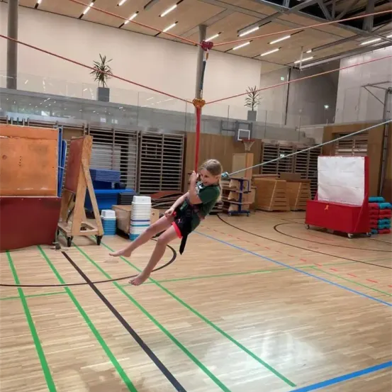 Ein junger Junge schwingt auf einer Schaukel in einer Turnhalle. Die Turnhalle hat Holzböden, Wände mit Topfpflanzen und verschiedenes Sportequipment.