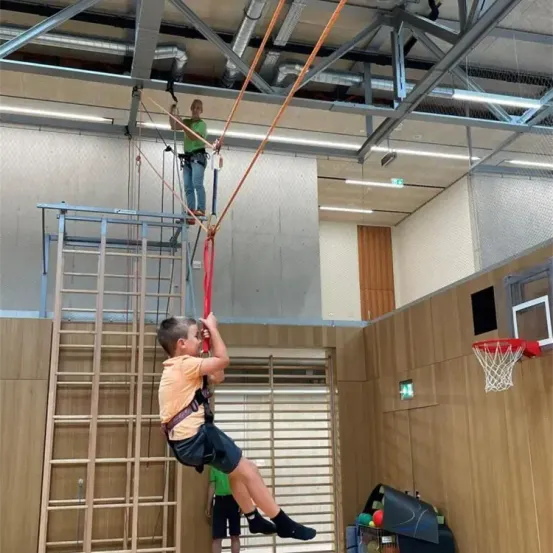 Zwei Kinder klettern auf einem Seil. Eines hängt, während das andere auf einer Plattform steht. Ein Basketballkorb ist an einer Wand befestigt.