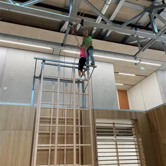 Zwei Personen klettern eine Leiter, die an einem Deckengurtsystem befestigt ist, wobei eine Frau einem Kind hilft.