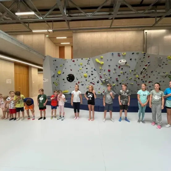 Eine Gruppe von Kindern steht vor einer Kletterwand mit verschiedenen farbigen Griffen und schaut nach oben und unterhält sich.
