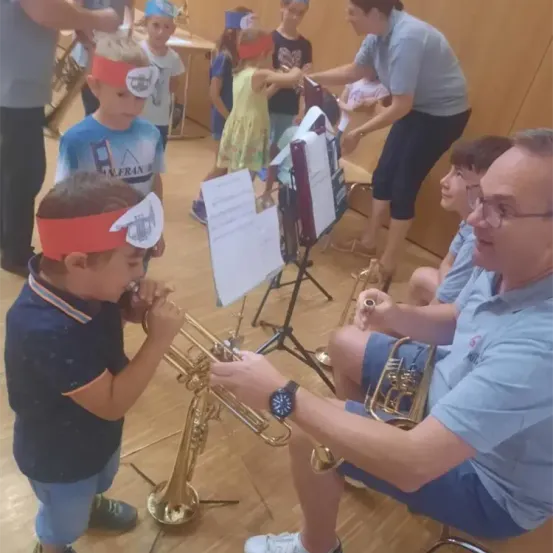 Eine Gruppe von Kindern spielt Trompeten mit Hilfe von Erwachsenen in einem Musikunterricht. Sie tragen Stirnbänder mit Trompetenbildern.