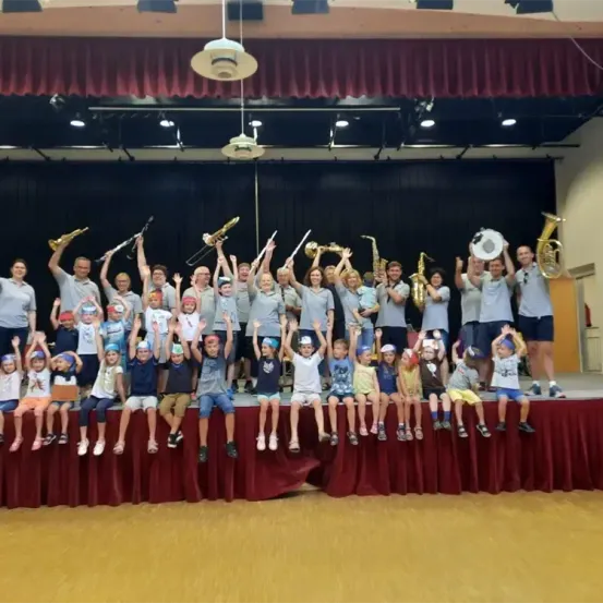 Eine Gruppe von Menschen auf der Bühne, die Musikinstrumente halten. Einige Erwachsene und Kinder. Roter Vorhang im Hintergrund.