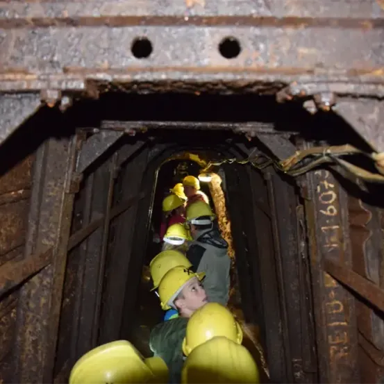 Eine Gruppe von Menschen in gelben Helmen kriecht durch einen dunklen, engen Tunnel. Der Tunnel ist alt und rostig, mit etwas Text und Zahlen an den Wänden.