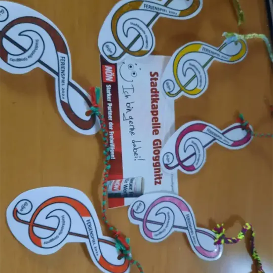 Eine dekorative Holzoberfläche zeigt farbenfrohe Musiknoten-Aufkleber, eine rote und weiße Karte mit 'Stadtkapelle Glognitz' und Schnüre, die die Aufkleber verbinden, möglicherweise für eine festliche Veranstaltung.