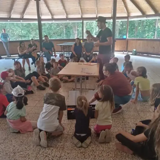 Ein Mann in einem Hut steht an einem Tisch und spricht eine Gruppe von Kindern an, die im Kreis sitzen. Erwachsene sitzen hinter den Kindern. Eine Frau steht in der Nähe des Tisches.