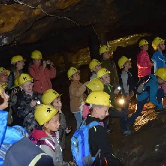 Eine Gruppe von Kindern erkundet eine Höhle, trägt gelbe Helme und einige haben Rucksäcke. Einige machen Fotos mit ihren Handys.
