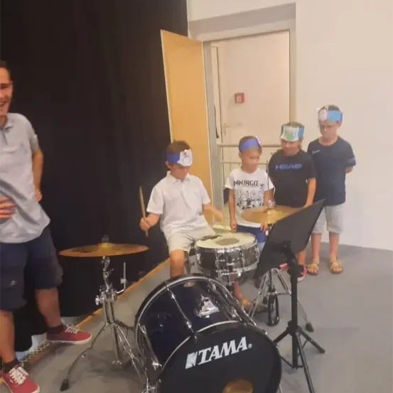 Ein Mann und mehrere Jungen spielen Trommeln mit verbundenen Augen. Sie befinden sich in einem Raum mit einem schwarzen Vorhang.