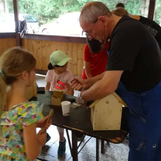 Ein älterer Mann unterrichtet zwei junge Mädchen im Bau eines Vogelhauses. Sie stehen um einen Tisch herum, auf dem ein hölzerne Vogelhaus und Werkzeuge liegen.