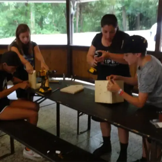 Vier Personen, zwei Frauen und zwei Männer, sind mit Holzarbeiten beschäftigt. Sie benutzen Bohrer und Werkzeuge an einer Holzstruktur auf einem Tisch. Draußen ist ein Auto geparkt.