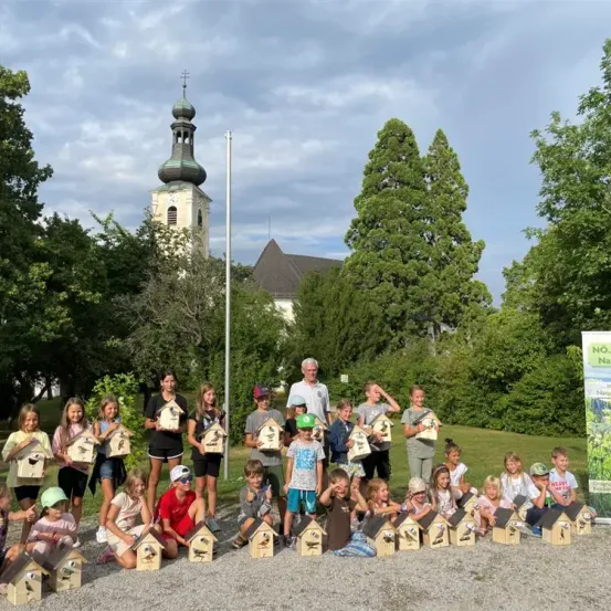 Eine Gruppe von Kindern und Erwachsenen posiert für ein Foto im Freien mit kleinen Vogelhäusern, in der Nähe einer Kirche und Bäumen.