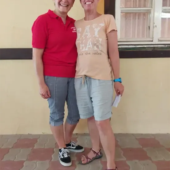 Zwei Frauen stehen nebeneinander und lächeln, eine in einem roten Poloshirt und die andere in einem beigen T-Shirt mit weißem Text. Sie stehen draußen in der Nähe eines Fensters mit einem gefliesten Boden.