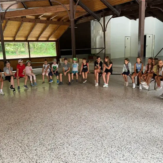 Eine Gruppe von Kindern sitzt in einem Kreis in einem Saal und trägt verschiedene Kleidungsstücke. Der Saal hat Holzbalken und einen offenen Raum.