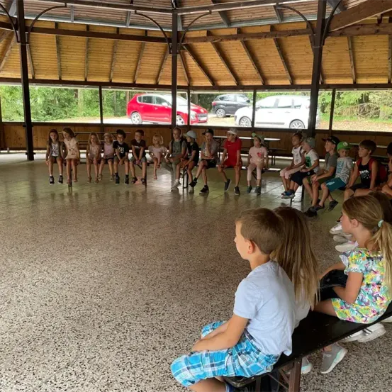 Eine Gruppe von Kindern sitzt im Kreis in einem Pavillon, wobei Autos durch die Fenster sichtbar sind. Die Kinder tragen Freizeitkleidung und scheinen jemandem zuzuhören.