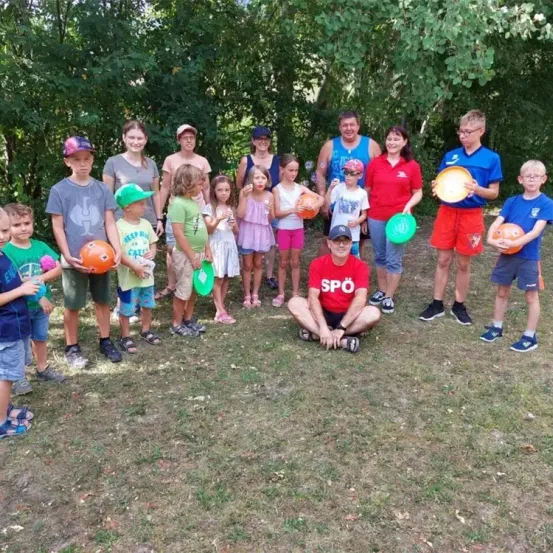 Eine Gruppe von Kindern und Erwachsenen steht im Freien im Kreis. Einige halten Bälle und Frisbees. Der Mann in der Mitte sitzt auf dem Boden. Hinter ihnen stehen Bäume.
