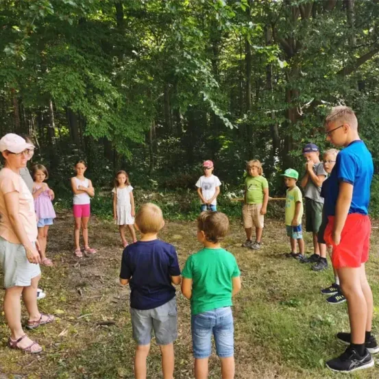 Eine Gruppe von Kindern und Erwachsenen steht in einem Wald. Sie tragen alle Freizeitkleidung. Der Mann vorne trägt eine Brille und ein blaues Hemd.