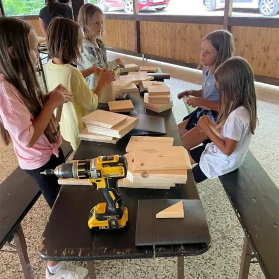 Eine Gruppe junger Mädchen arbeitet an einem Holzprojekt an einem Tisch und benutzt eine Bohrmaschine. Verschiedene Holzstücke sind auf dem Tisch verstreut. Sie sitzen auf Bänken in einer Außenwerkstatt.