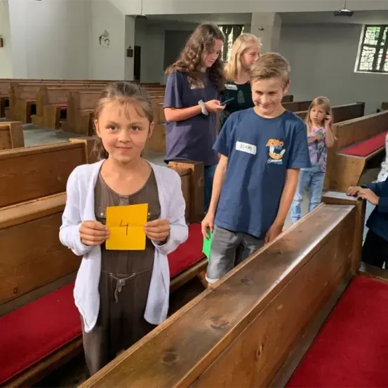 Eine Gruppe von Kindern ist in einer Kirche, ein Mädchen hält ein gelbes Papier mit der Nummer 4, ein anderer Junge lächelt. Andere Kinder und Erwachsene sind anwesend.