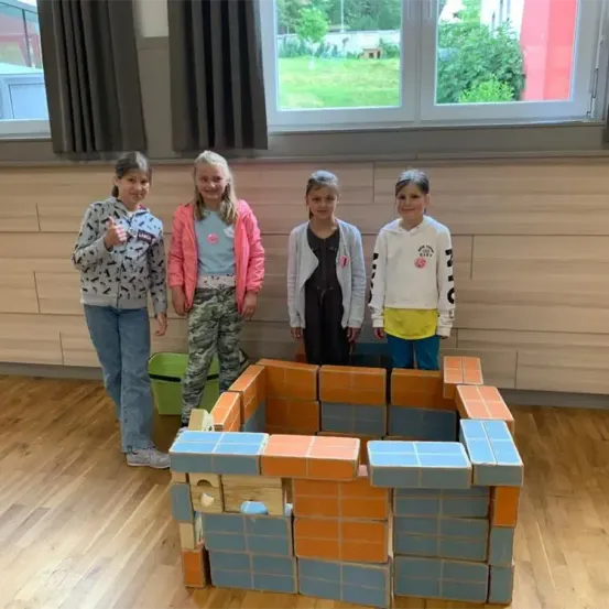 Vier junge Mädchen stehen neben einer Holzziegelstruktur in einem Raum. Die Struktur ist orange und blau. Vor dem Fenster befindet sich ein Rasenbereich.