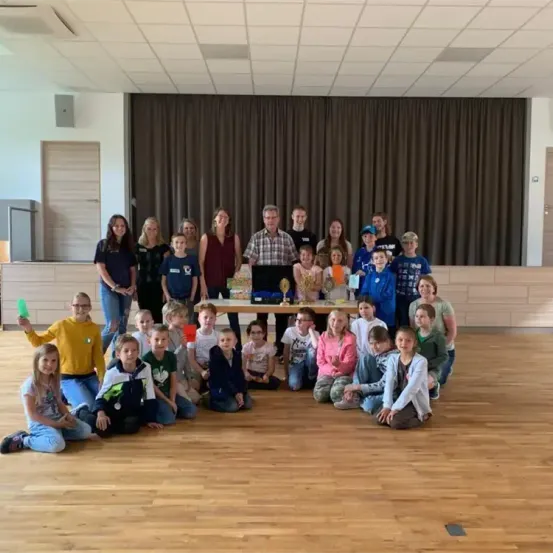 Gruppenfoto von Kindern, Lehrern und Erwachsenen in einem Raum mit Holzböden und einer Bühne. Einige Kinder halten Pokale.