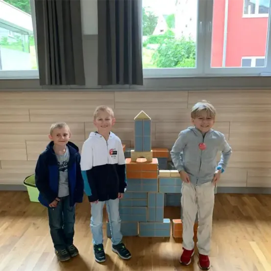 Drei junge Jungen stehen vor einer blauen und orangefarbenen Ziegelstruktur, lächeln und posieren für ein Foto in einem Raum mit Holzböden und großen Fenstern.