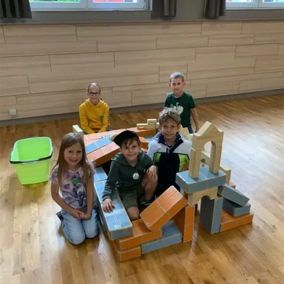 Fünf Kinder spielen mit Holzklötzen in einem Raum. Sie sitzen und stehen um eine große Struktur aus Ziegeln herum. Ein grüner Eimer ist links.