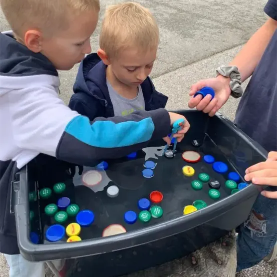 Drei junge Jungen spielen mit Wasser und Plastikflaschenverschlüssen in einer großen Plastikwanne. Ein Junge trägt eine weiße und blaue Jacke. Ein anderer Junge trägt eine blaue Jacke. Ein Erwachsener hält ein blaues Objekt in der Hand.