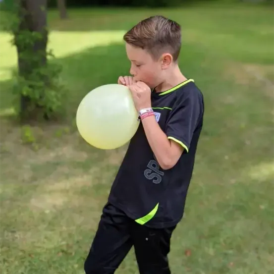 Ein junger Junge bläst einen gelben Ballon auf einem Grasfeld auf. Er trägt ein schwarzes Trikot mit den Buchstaben SP.