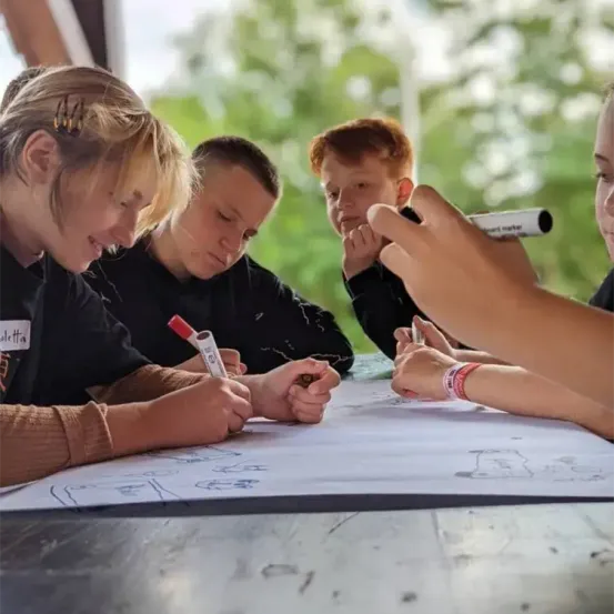 Fünf Kinder sitzen um einen Tisch herum, jeder hält Marker und zeichnet auf einem großen Blatt Papier. Ein Kind trägt einen Namensschild mit der Aufschrift 'Valetta'. Bäume sind im Hintergrund zu sehen.