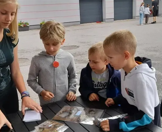 Eine Frau mit blonden Haaren unterrichtet drei Jungen an einem Tisch. Sie betrachten einige Plastiktüten. Im Hintergrund befinden sich Gebäude und Menschen.