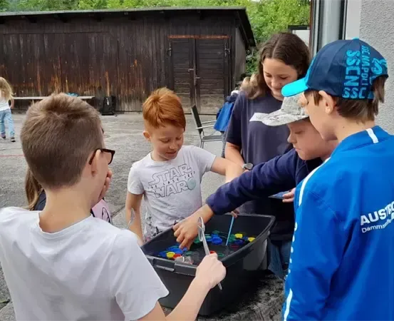 Mehrere Kinder spielen mit Spielzeug in einem Behälter im Freien. Sie sind draußen in der Nähe eines Gebäudes und anderer Kinder.