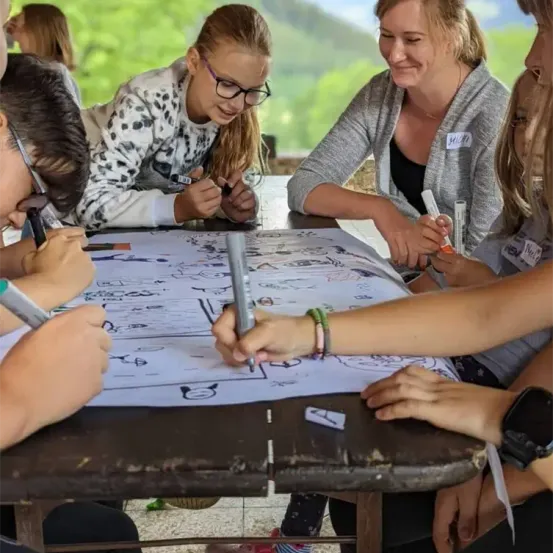 Eine Gruppe von Menschen, darunter Kinder, versammelt sich um einen Tisch und ist in einer Zeichenaktivität verstrickt. Sie halten Marker und scheinen ein großes Bild auf einem Blatt Papier zu erstellen.