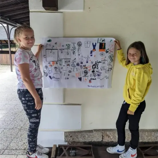 Zwei junge Mädchen halten ein großes Blatt Papier mit verschiedenen Zeichnungen hoch. Sie stehen draußen, und die Wand hinter ihnen ist gelb gestrichen. Das Mädchen auf der linken Seite trägt ein weißes Shirt und schwarze Hose mit Schriftzügen. Das Mädchen auf der rechten Seite trägt eine gelbe Hoodie und schwarze Hose.