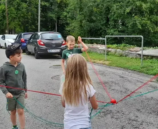 Drei Kinder spielen auf der Straße mit roten und grünen Seilen. Ein graues Auto ist in der Nähe geparkt.