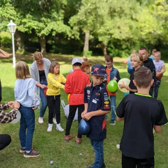 Eine Gruppe von Kindern und Erwachsenen versammelt sich in einem Park und hält Luftballons. Einige tragen Mützen und Turnschuhe. Der Junge in der Mitte hält einen blauen Helm.