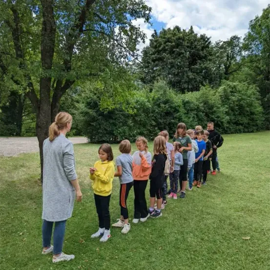 Eine Frau steht in einem Park mit Kindern, einige in Turnschuhen, in einer Reihe vor ihr. Sie sind von üppiger Grünfläche umgeben.