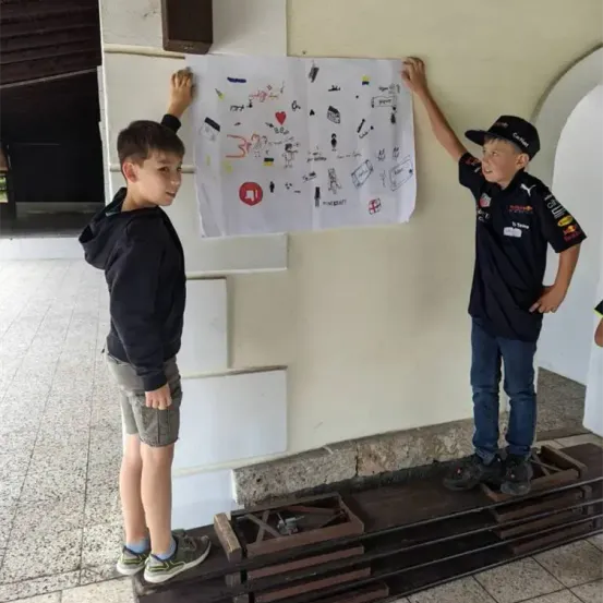 Zwei Jungen stehen neben einer Wand und halten ein weißes Poster mit Zeichnungen und Text. Sie tragen Freizeitkleidung und Sneakers. Der Junge auf der linken Seite trägt eine Kapuzenjacke, während der Junge auf der rechten Seite eine Kappe und ein Shirt mit Logos trägt.