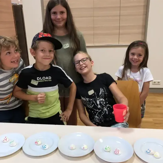 Vier Kinder, drei Jungen und ein Mädchen, sitzen um einen Tisch herum, auf dem kleine Teller mit lächelnden Gesichtern stehen. Zwei der Jungen tragen eine Brille und eine Kappe, während die Mädchen lächeln und hinter dem Tisch stehen.