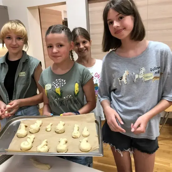 Vier Mädchen in einer Küche, von denen eines eine Schale mit ungebackenen Keksformen hält. Das Mädchen mit der Schale trägt ein graues T-Shirt mit Blumen- und Maisdesign.