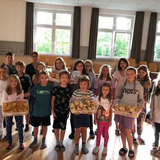 Eine Gruppe von Kindern und Erwachsenen steht in einem Raum und hält Tabletts mit Essen. Sie tragen Freizeitkleidung. Hinter ihnen stehen zwei Erwachsene in der Nähe der Fenster.